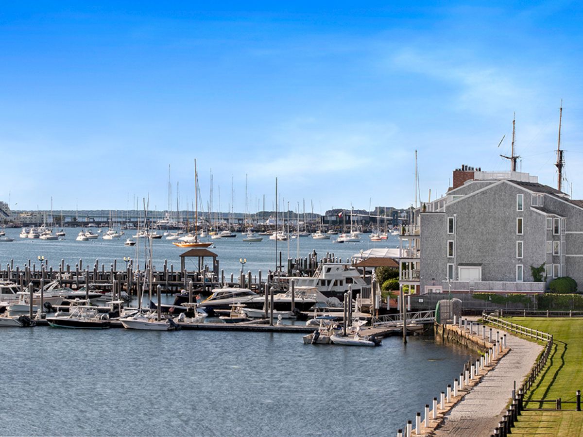 The image shows a marina with multiple boats docked, a waterfront building, and calm water under a clear blue sky.