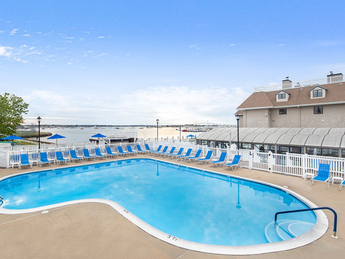 Outdoor pool with blue lounge chairs, overlooking a waterfront with a view of boats. There's a building and white fencing around the area.