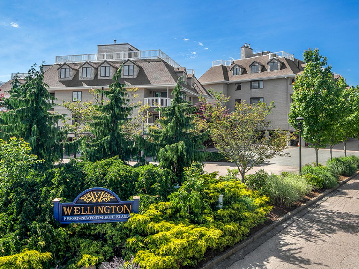 The image shows a residential building with a sign saying "Wellington," surrounded by lush greenery and blue skies.