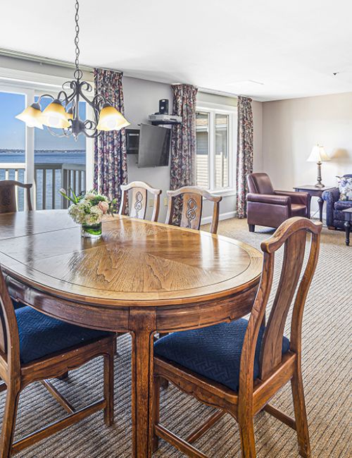 The image shows a dining and living area with a wooden table, chairs, couches, and a view of the ocean through large windows.