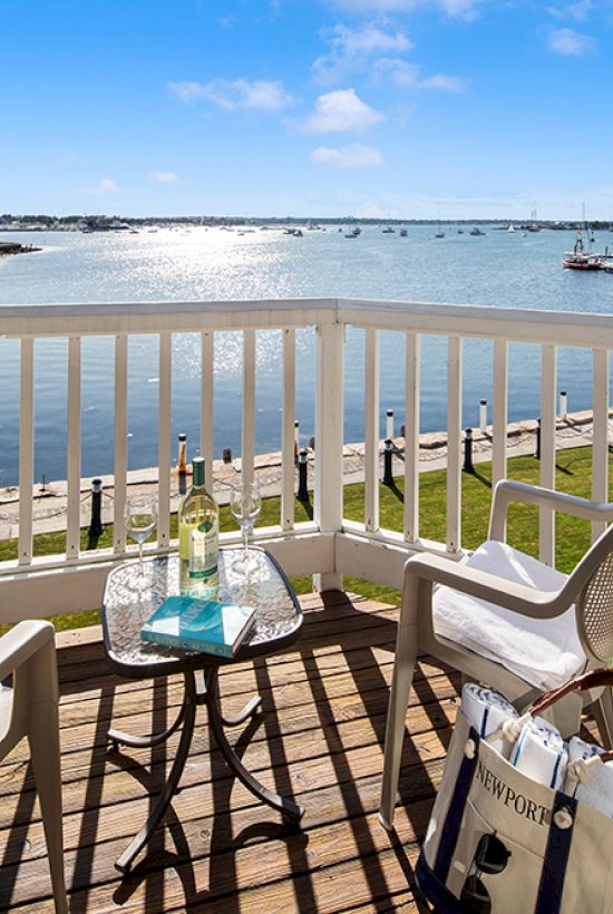 A balcony with two chairs and a table overlooks a scenic view of water, boats, and green landscape, under a clear blue sky.