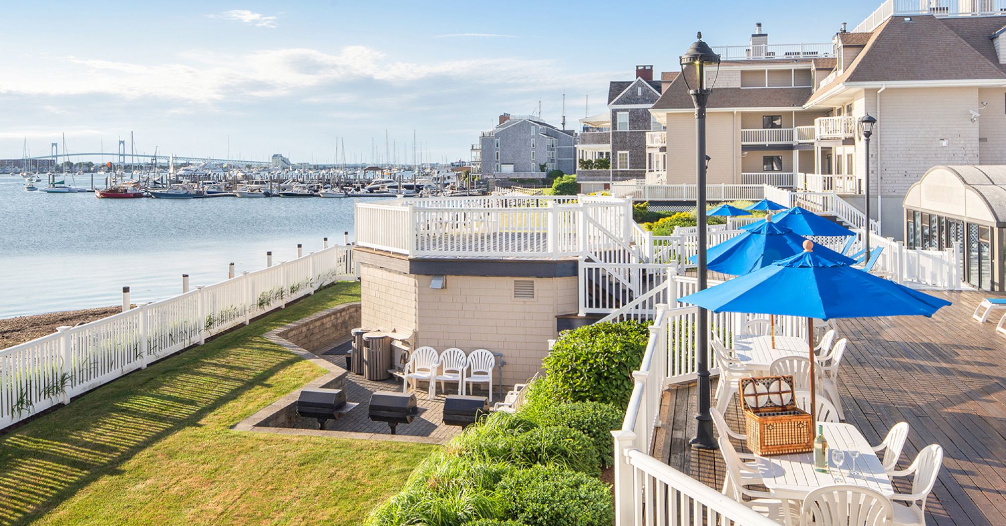 A waterfront view with a wooden deck, blue umbrellas, white chairs, and nearby marina boats.