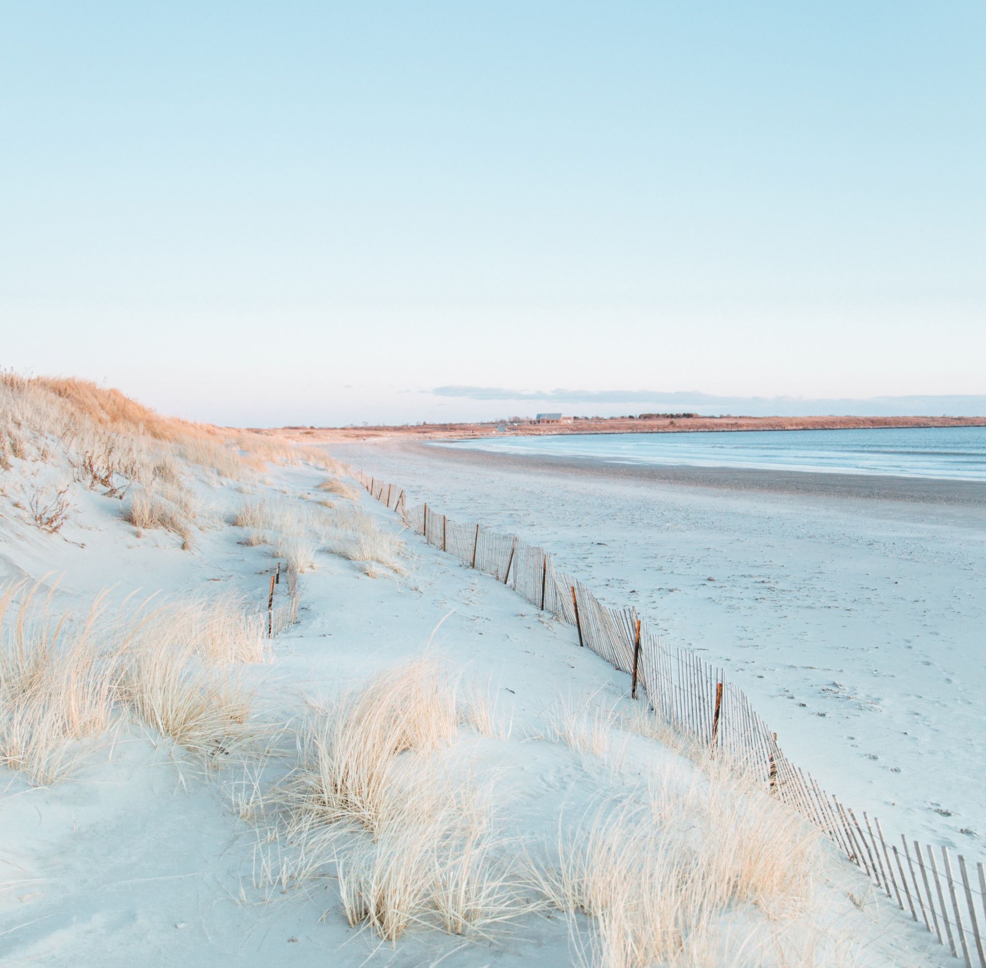 A serene beach with sand dunes, sparse grass, and a wooden fence extending toward the horizon under a clear blue sky.