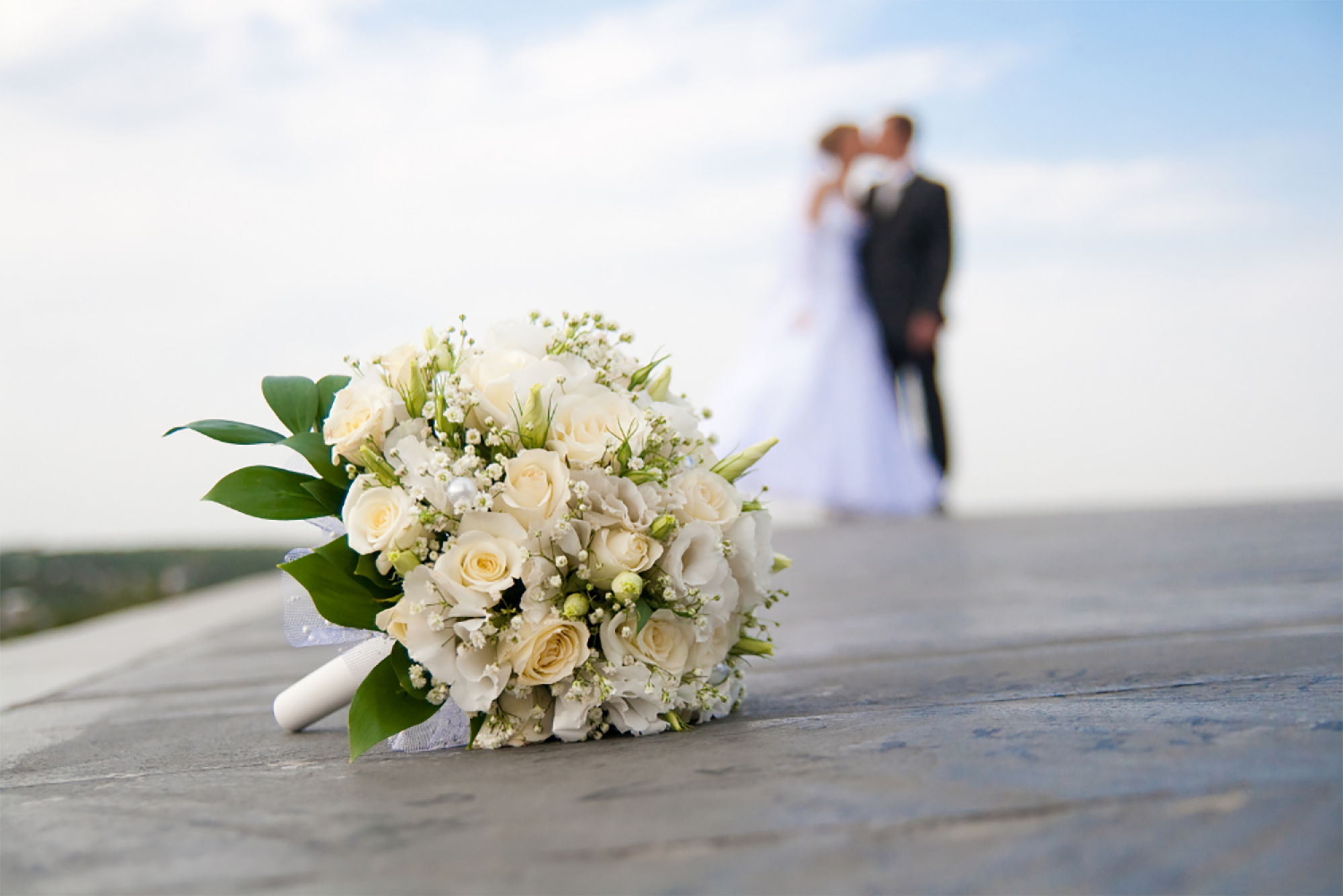 A bouquet of white roses and greenery in focus; a blurred couple in wedding attire stands in the background against a cloudy sky.