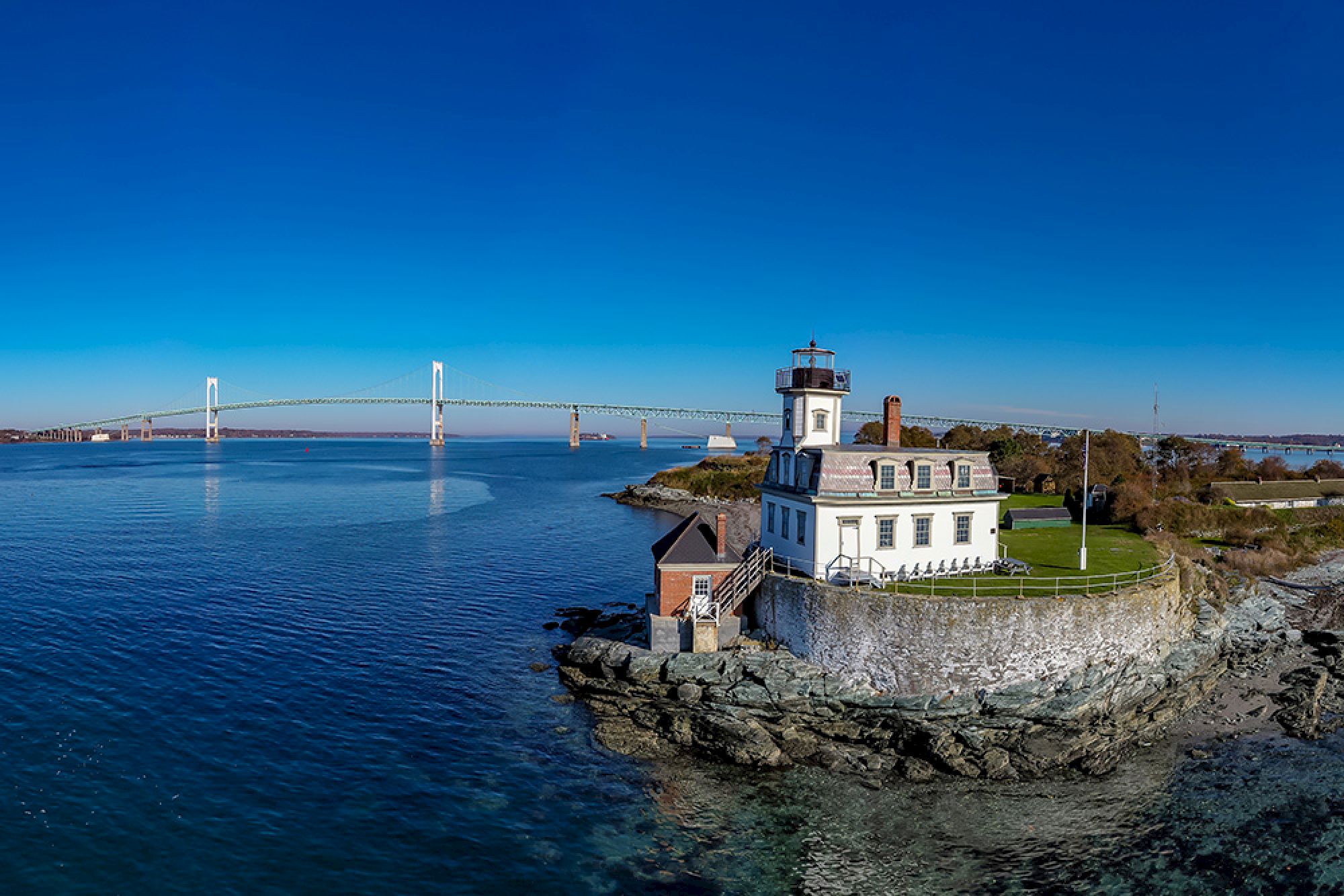 A lighthouse sits on a rocky coast with a nearby bridge in the background, surrounded by calm blue water and clear skies.