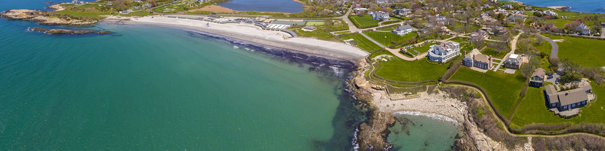 This image shows a coastal landscape with turquoise waters, rocky shores, sandy beaches, and homes on green fields under a clear blue sky.