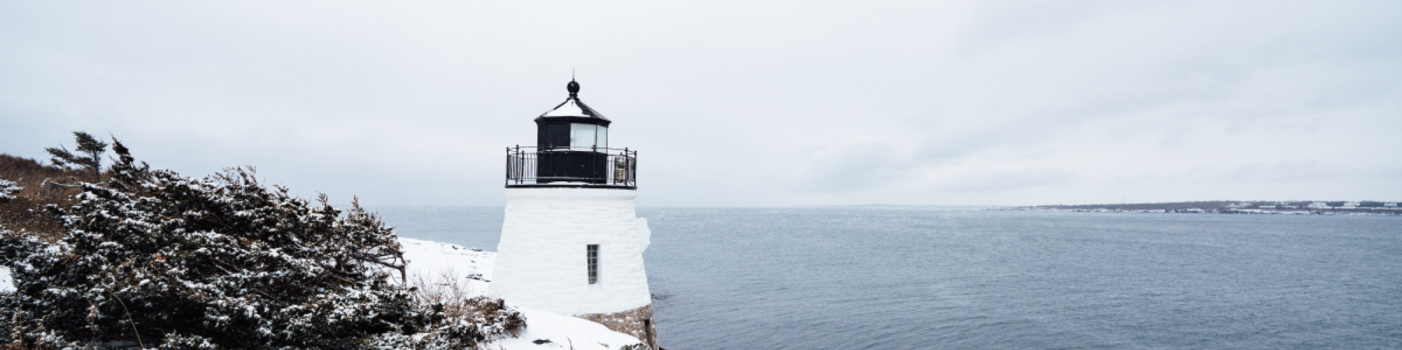 A snowy coastal scene with a white lighthouse overlooking the ocean, surrounded by shrubs and grass.