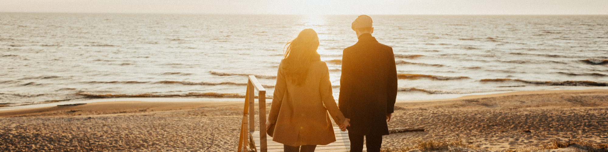 A couple is walking hand in hand towards the beach, with the sun setting over the horizon and a wooden boardwalk beneath their feet.