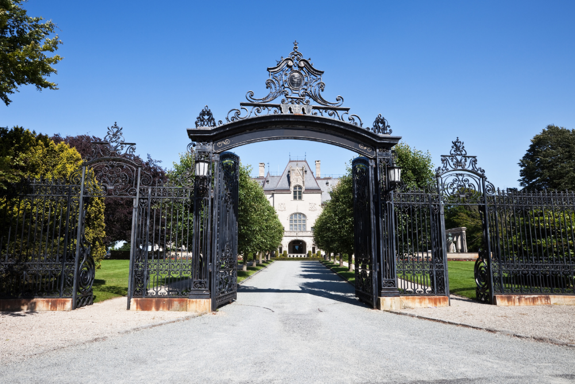 An ornate iron gate opens to a grand mansion with a long driveway, flanked by trees under a clear blue sky.