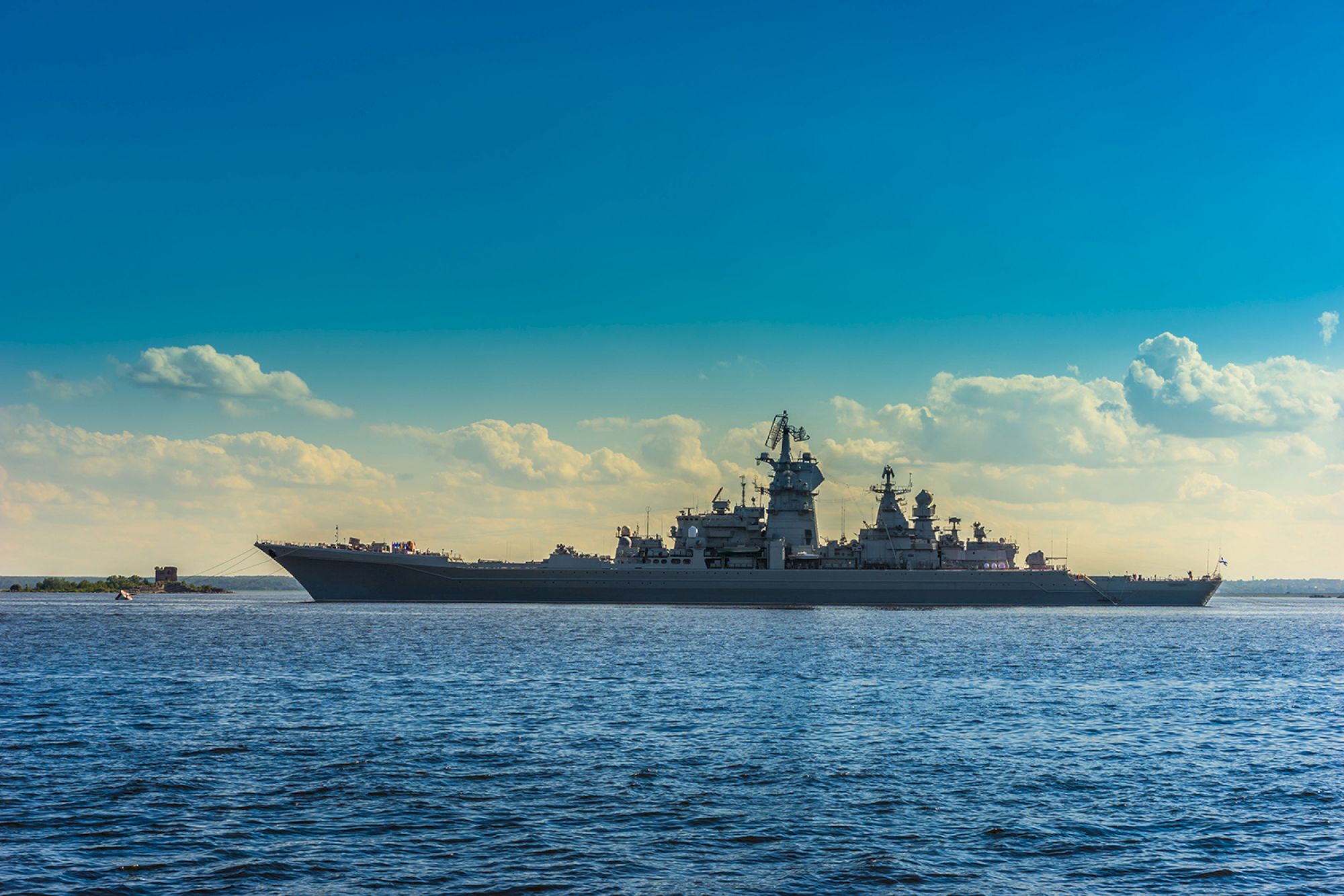 A large battleship sails on calm waters under a clear blue sky with scattered clouds in the background.