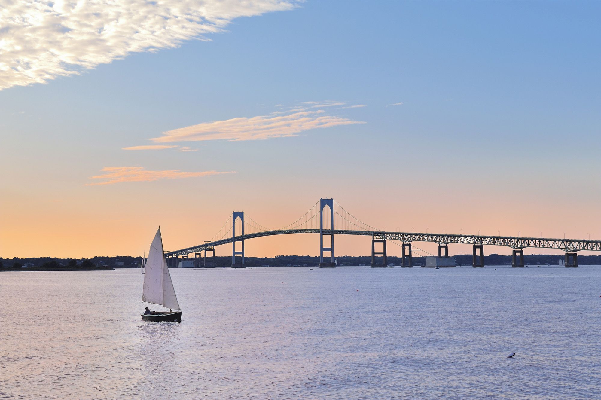 A sailboat on calm water with a bridge in the background during a sunset, with a partly cloudy sky above and warm colors reflecting.