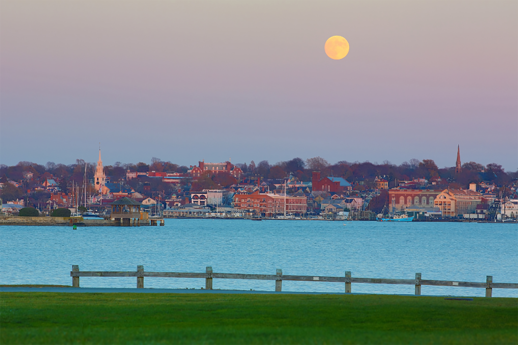 A scenic view of a waterfront town at dusk, with a full moon in the sky and a fence in the foreground.