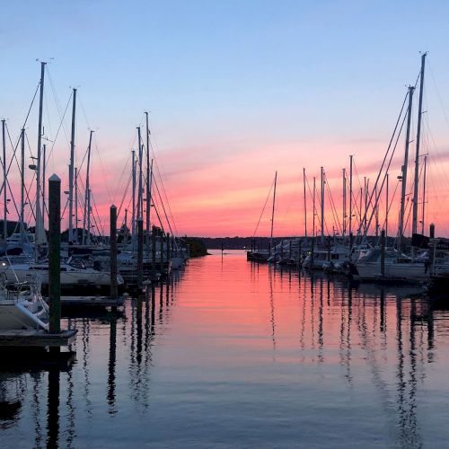 A marina at sunset with boats docked on both sides of a calm waterway, framed by a colorful sky.