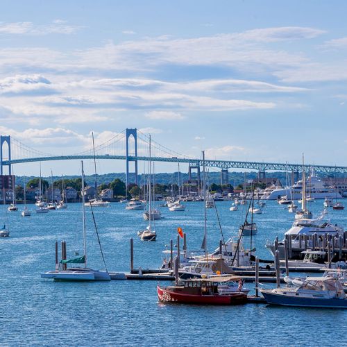 The image shows a scenic view of a marina with sailboats and yachts, and a bridge in the background under a partly cloudy sky.