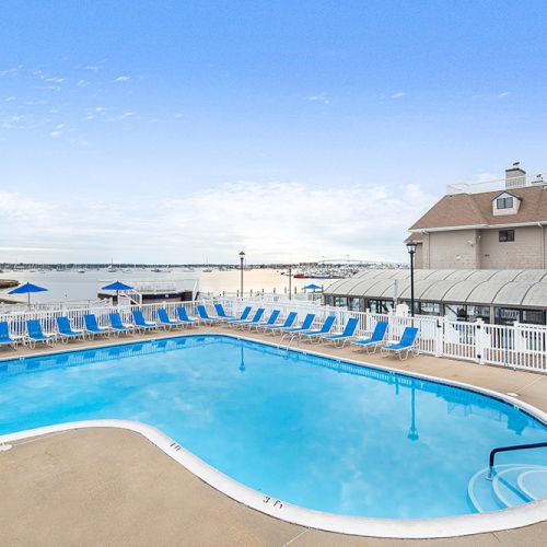 A pool with lounge chairs, a view of the sea, and a building nearby under a clear blue sky.