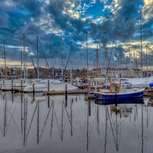 A marina with boats and sailboats docked, reflecting in calm water under a dramatic, cloudy sky with hints of blue.