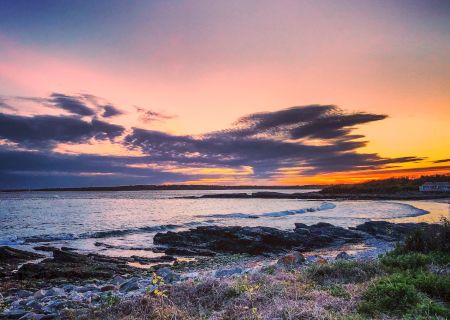 A scenic coastal sunset with colorful skies, rocky shoreline, and calm sea waves create a serene and picturesque view.