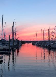 A marina at sunset with boats docked and masts silhouetted against a colorful sky, reflecting on the calm water, creating a serene scene.