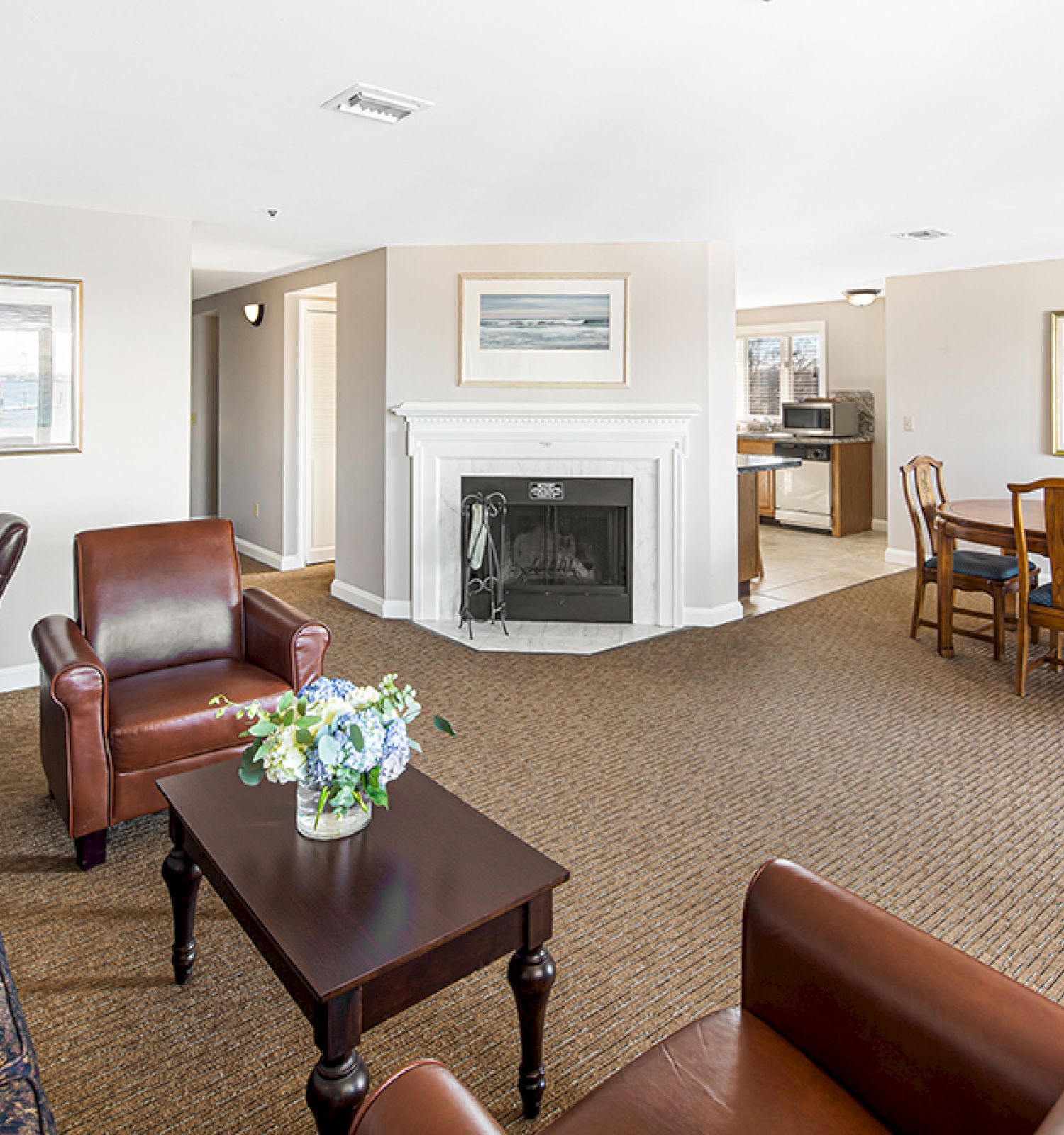 The image shows a spacious living room with a fireplace, armchairs, sofa, dining table, and decorative plants and paintings on the walls.