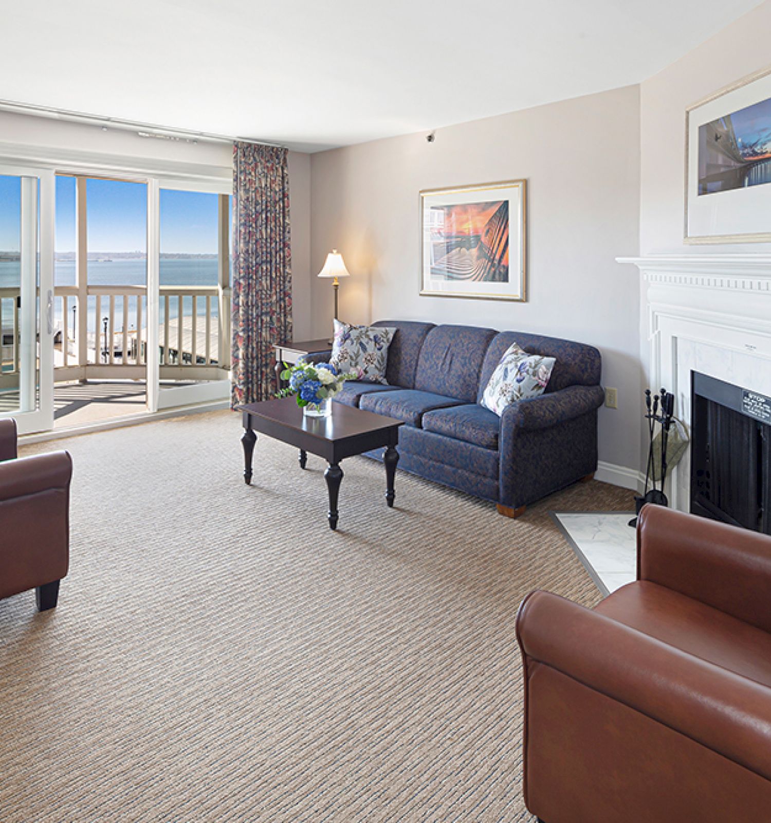 A cozy living room with two leather chairs, a couch, a coffee table, and a fireplace, overlooking a balcony with an ocean view.