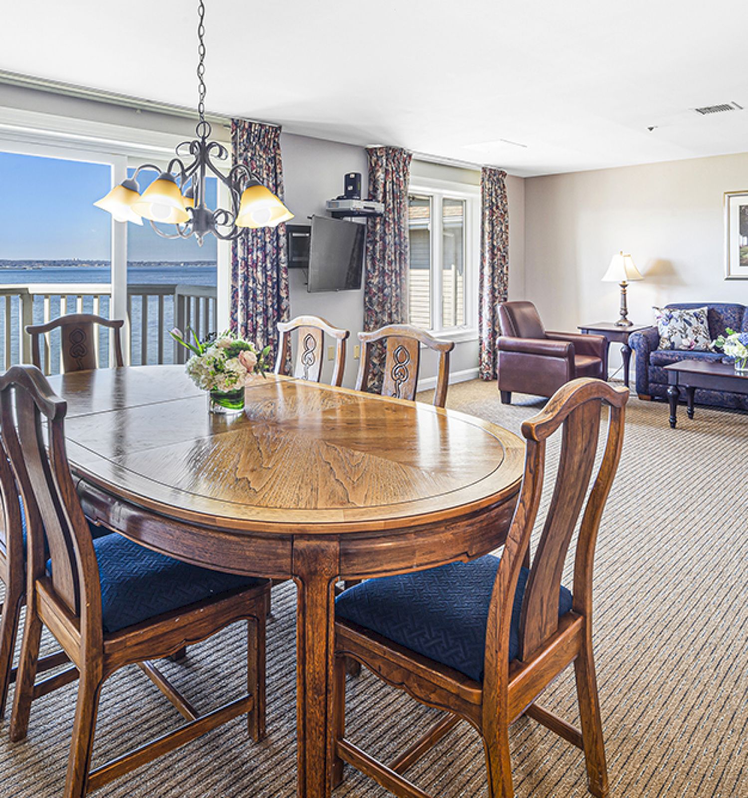 The image shows a dining and living area with a wooden table, chairs, sofas, a lamp, and large windows overlooking a body of water.