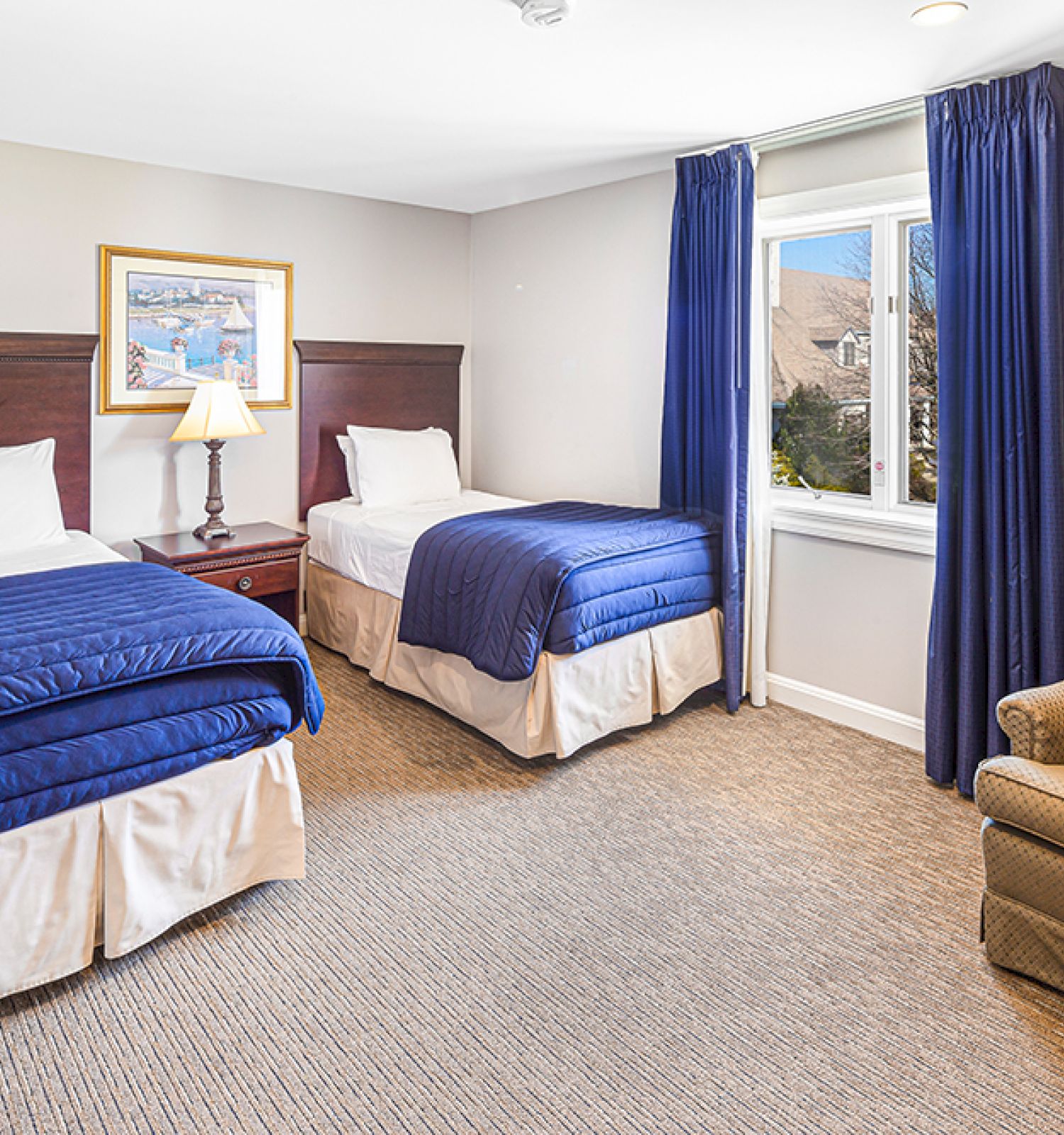 The image shows a hotel room with two single beds, blue bedding, a nightstand with a lamp, a window, a mirror, and a brown sofa.