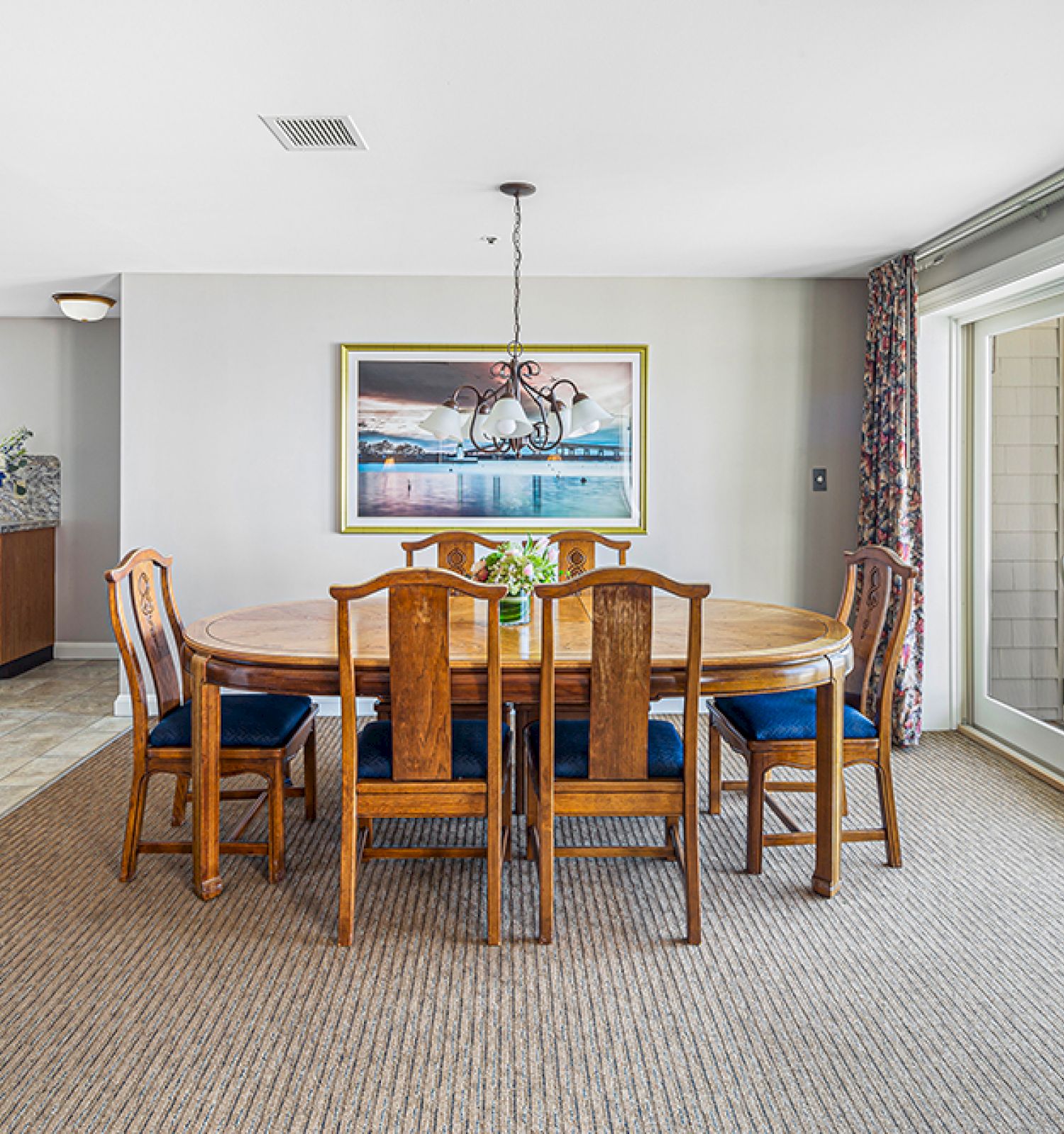 A dining room with a wooden table and six chairs, a light fixture, artwork on the wall, and a view through large windows to a balcony.