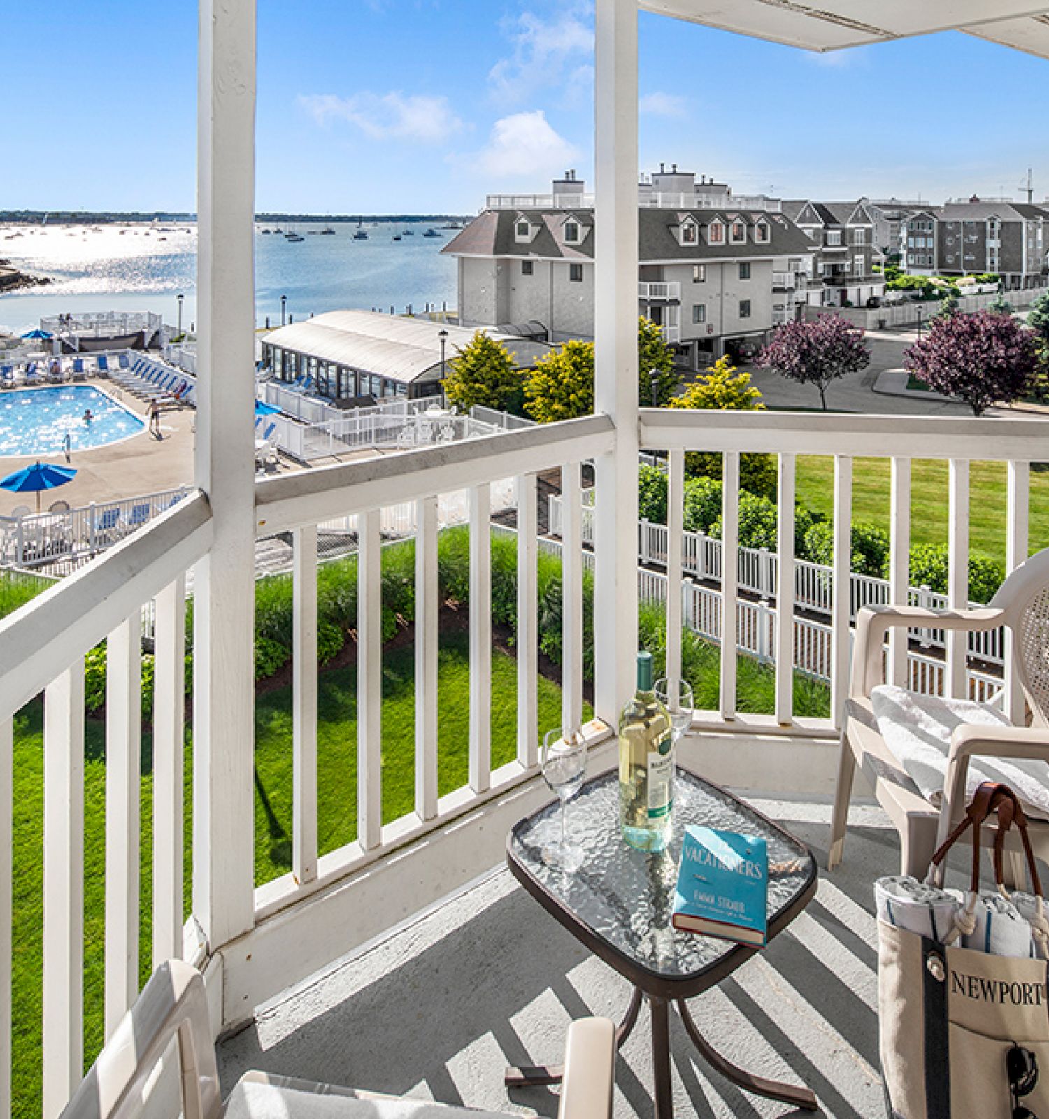 A seaside resort with a pool view, chairs on a deck, and buildings in the distance under a clear blue sky.