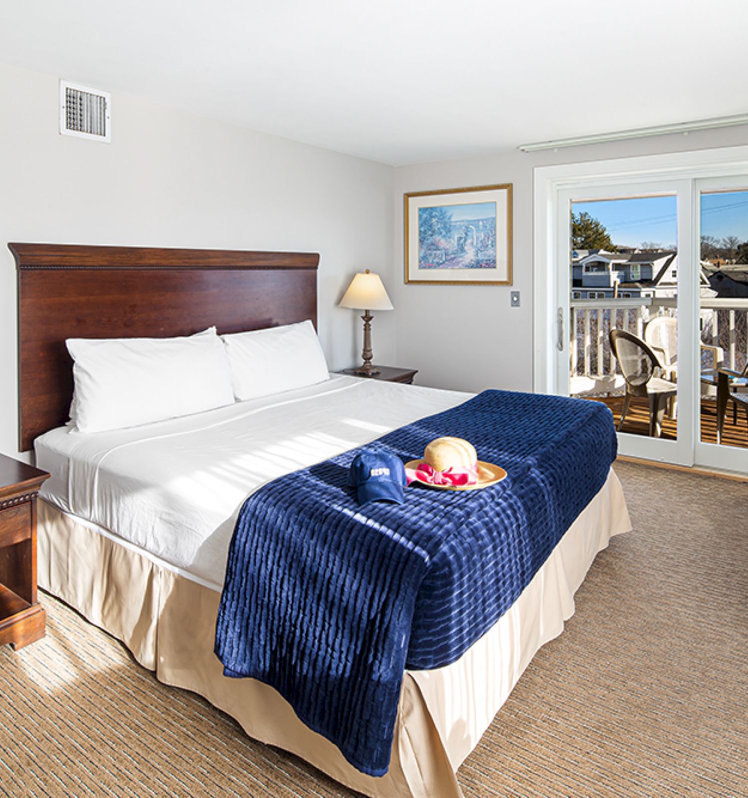 A cozy hotel room with a king-sized bed, blue accents, a side table, lamps, artwork, and a balcony with a view outside.