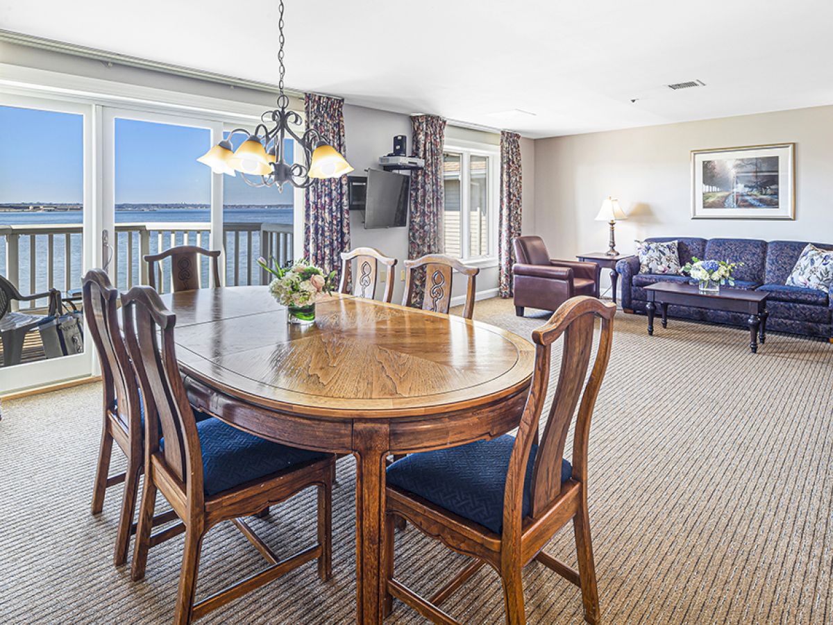 A spacious living and dining area with a large wooden table, chairs, couches, lamps, and sea view through large windows completes the setting.