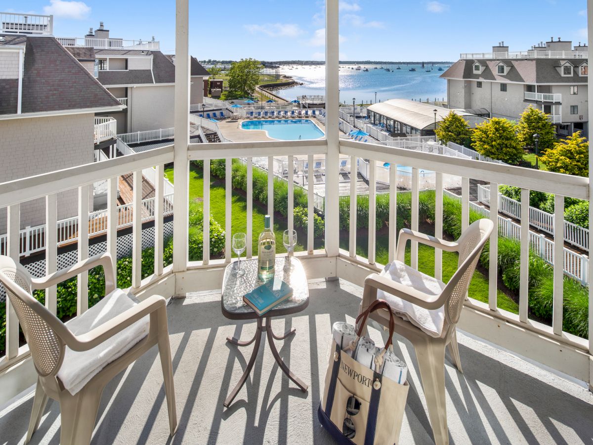 A balcony view with two chairs, a small table with drinks, and a tote bag overlooks a pool, greenery, and a scenic water area with buildings.