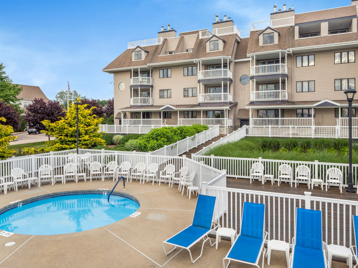 A residential building with a small fenced pool area surrounded by blue lounge chairs and greenery on a sunny day.