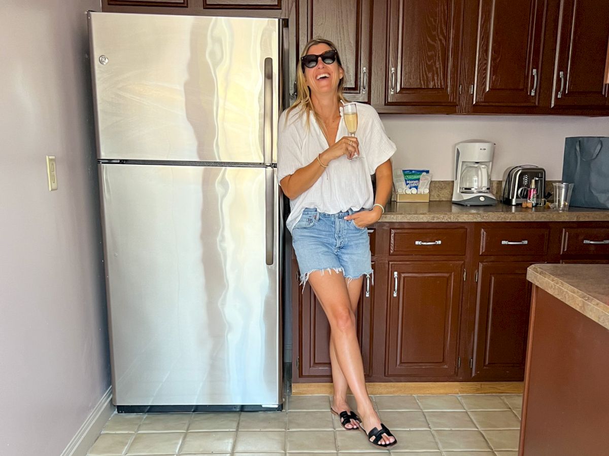 A person in a casual outfit stands in a kitchen, leaning against a fridge and holding a drink, with brown cabinets in the background.