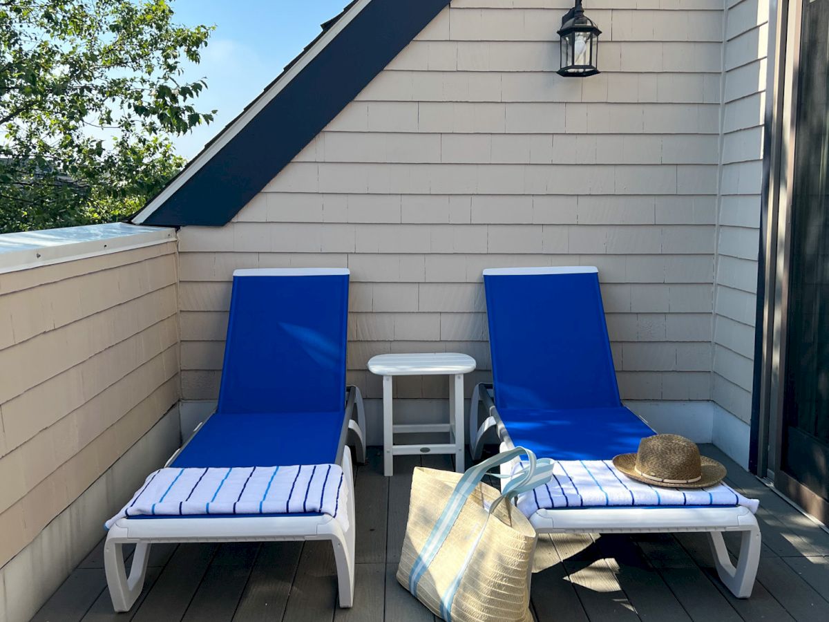 Two blue lounge chairs on a patio, with a small table, straw bag, and hat. There's a wall-mounted lamp above the chairs.