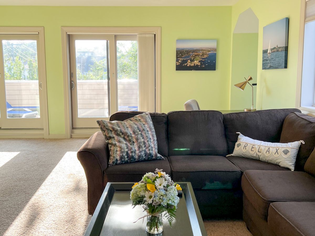 A bright living room with a brown sectional sofa, a coffee table with flowers, and wall art. Sunlight streams through glass doors.