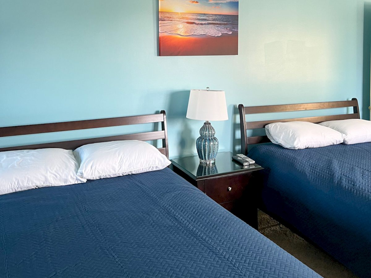 Two neatly made beds with blue bedspreads are in a room with a central nightstand and lamp, under a beach-themed picture.