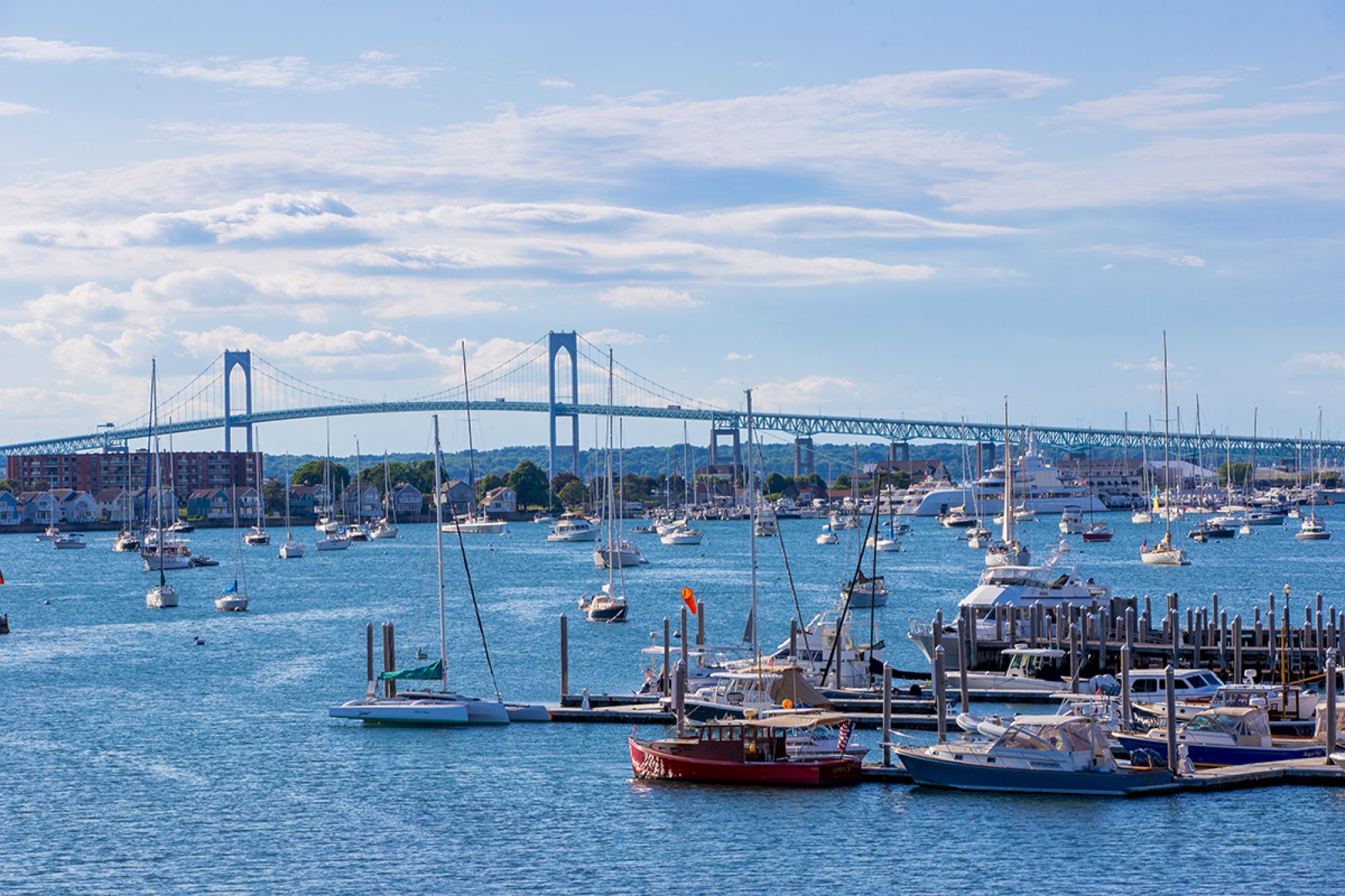 The image shows a harbor with docked boats and sailboats on water, a large bridge in the background, under a partly cloudy sky.