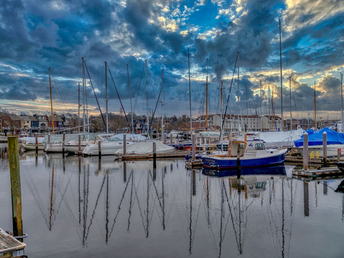 The image shows a marina with several sailboats docked, reflecting on the calm water under a dramatic, cloud-filled sky.