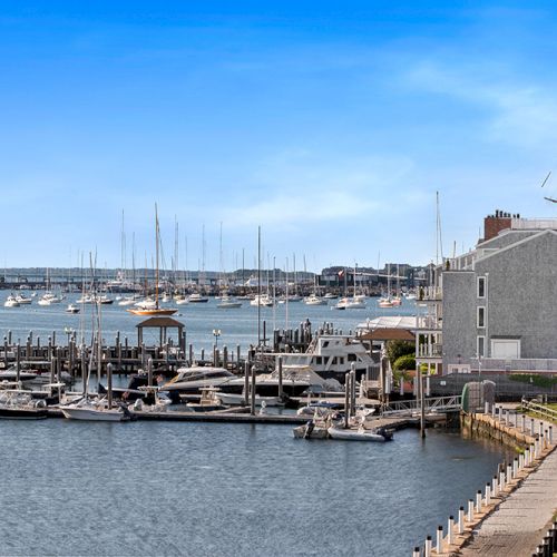 A marina with several boats docked, a large building, and a pathway alongside the water under a bright blue sky.