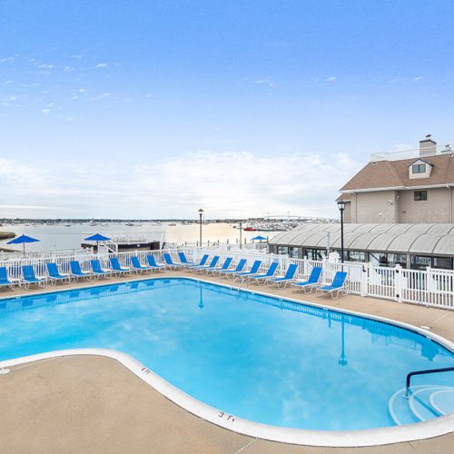 A resort pool with lounge chairs, a view of the beach and ocean in the background, and a building on the right under a clear blue sky.