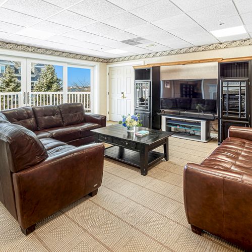 The image shows a living room with brown leather sofas, a black coffee table, and a large TV unit, offering a view of trees and buildings.