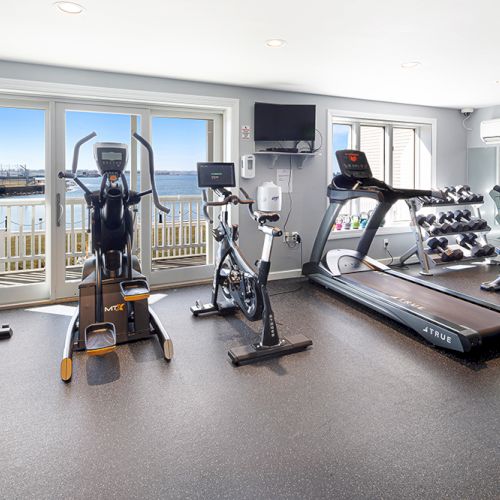 The image shows a gym with exercise equipment, including a treadmill, bike, and weights, facing large windows with a water view.