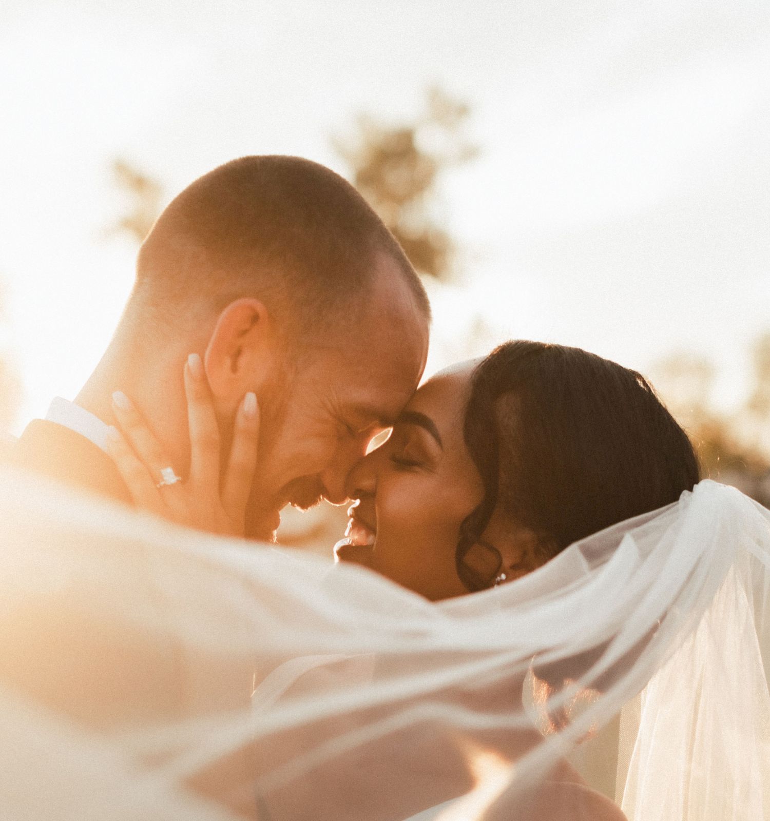 A couple is embracing and smiling on their wedding day, with soft sunlight and a veil flowing around them, creating a romantic scene.