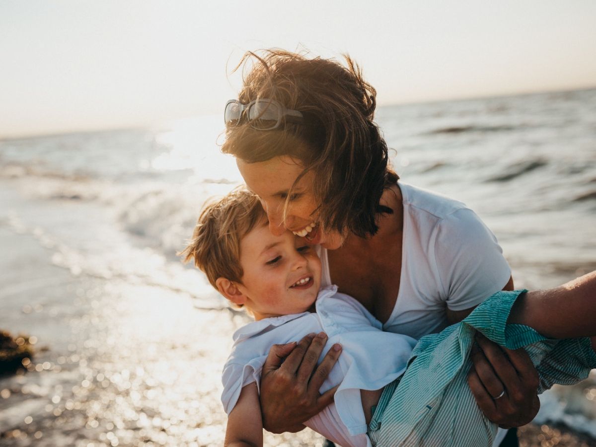 An adult joyfully holds a child by the ocean, both smiling, as the sun sets in the background, creating a warm and happy scene.