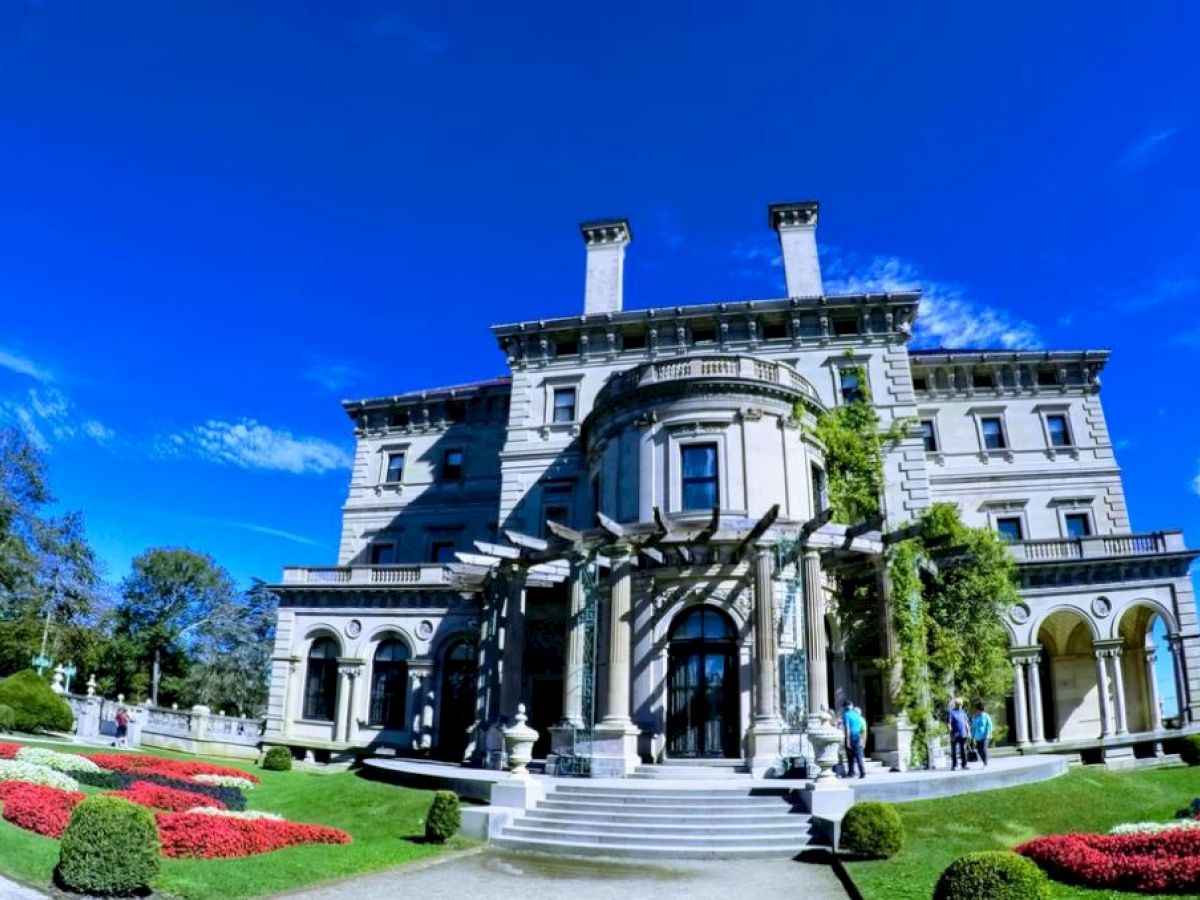 A grand mansion with a large entrance, gardens, and a bright blue sky above, surrounded by people and neatly trimmed hedges.