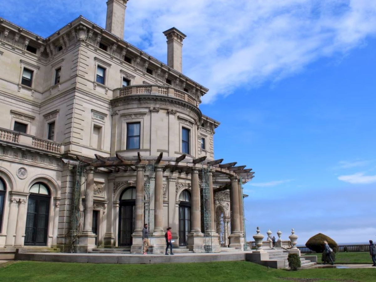 A large, ornate mansion with columns stands against a blue sky, with people walking around the grassy lawn in front.