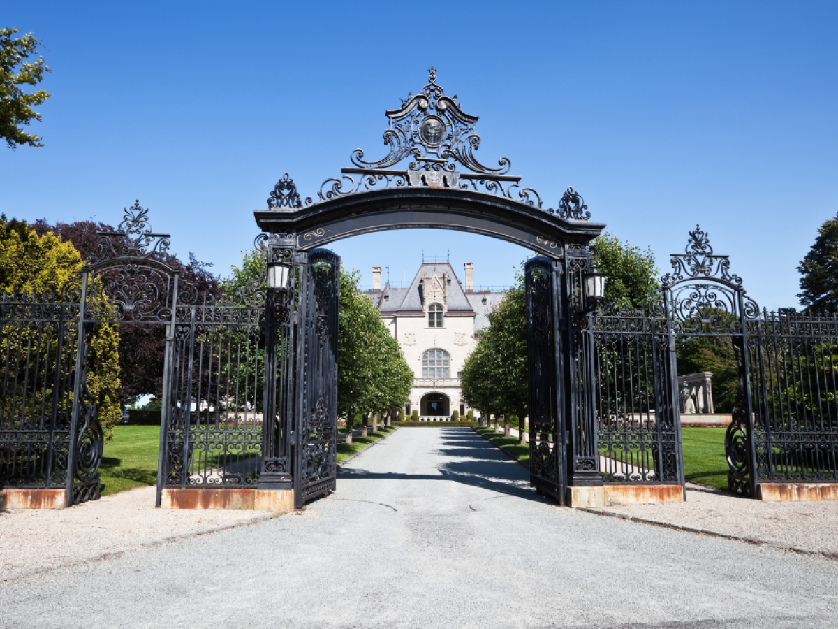 An ornate wrought iron gate opens to a pathway leading to a stately mansion surrounded by trees under a clear blue sky.