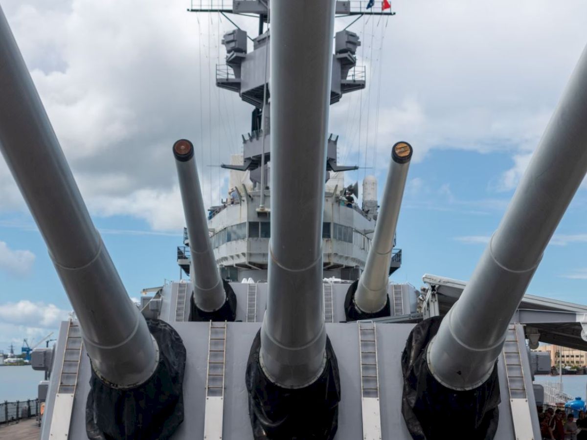 The image shows the large guns of a battleship, with a cloudy sky and waterfront visible in the background as people explore the deck.