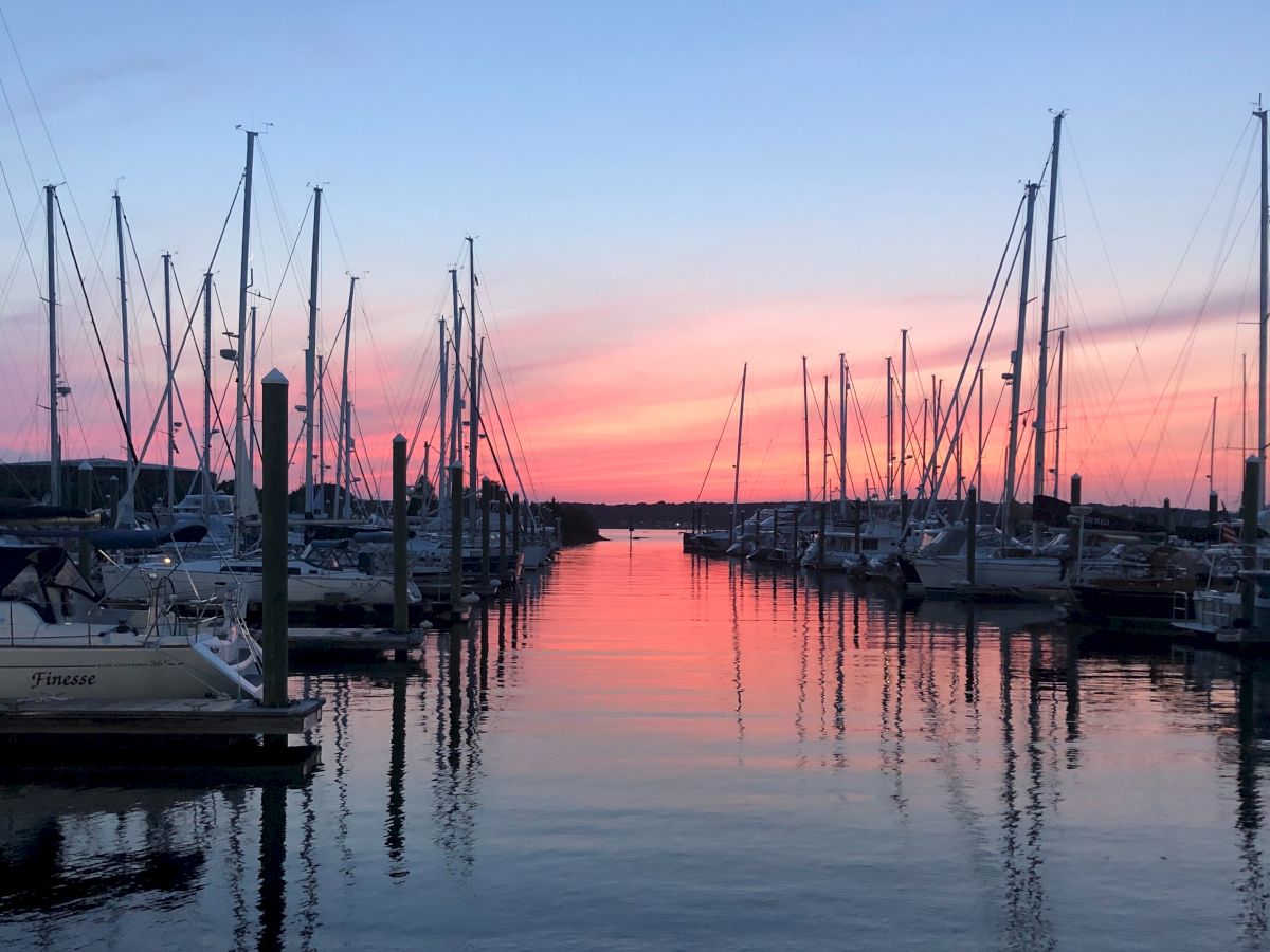 A marina scene with numerous boats docked, silhouetted by a vibrant sunset or sunrise over calm waters.