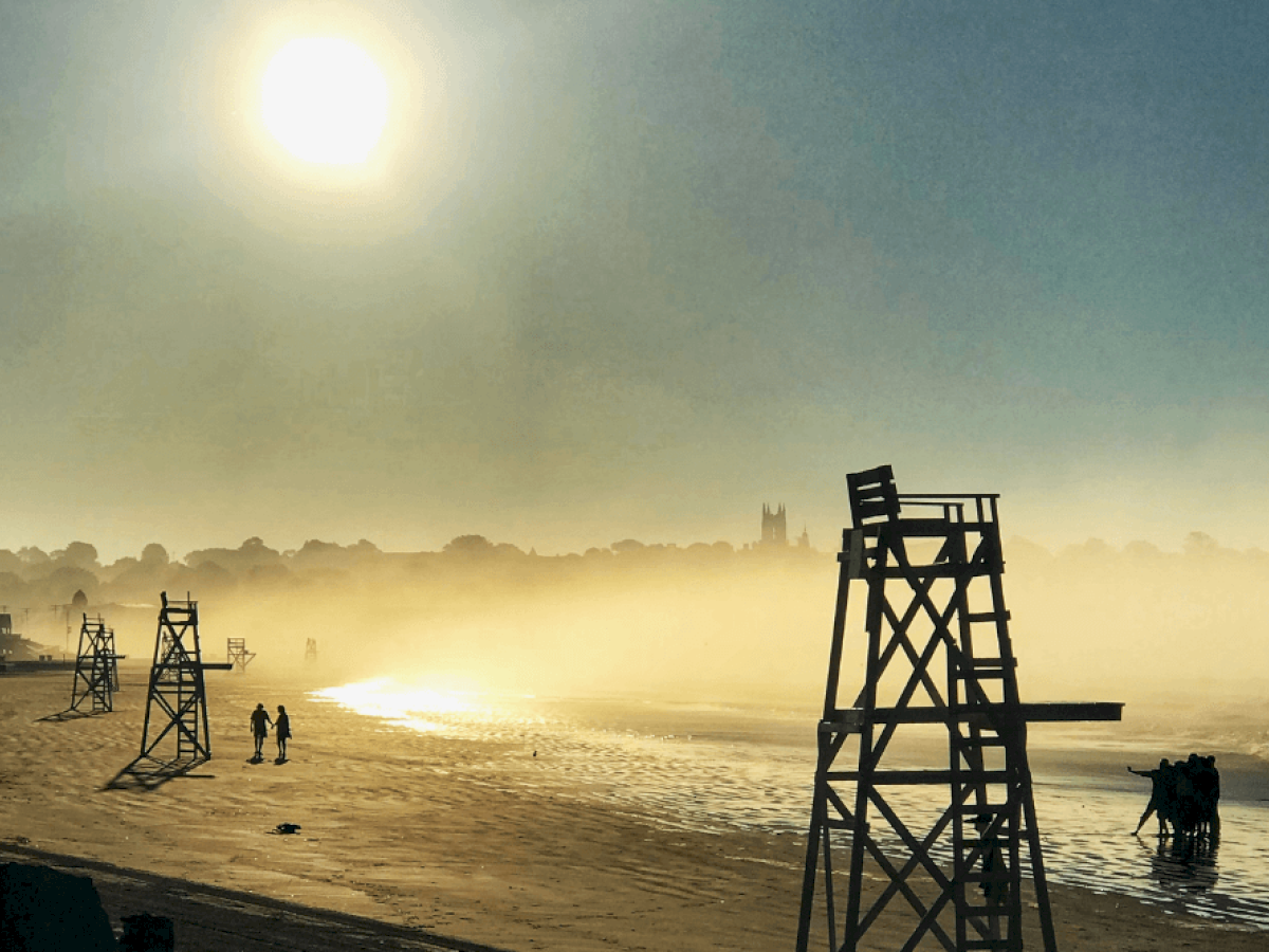 A foggy beach scene with lifeguard chairs, silhouettes of people, and a bright sun overhead.